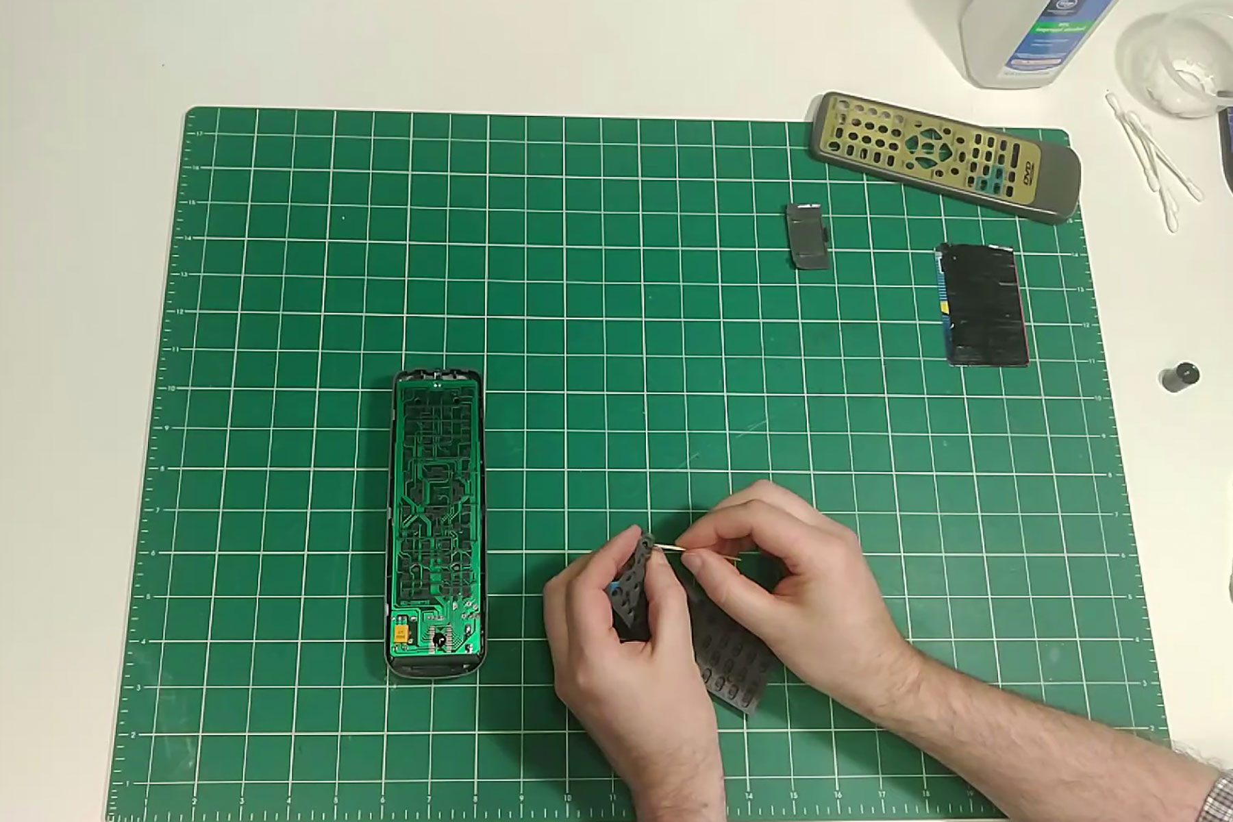 A person disassembles a device, holding a component over a green cutting mat, with various parts and cleaning supplies nearby.