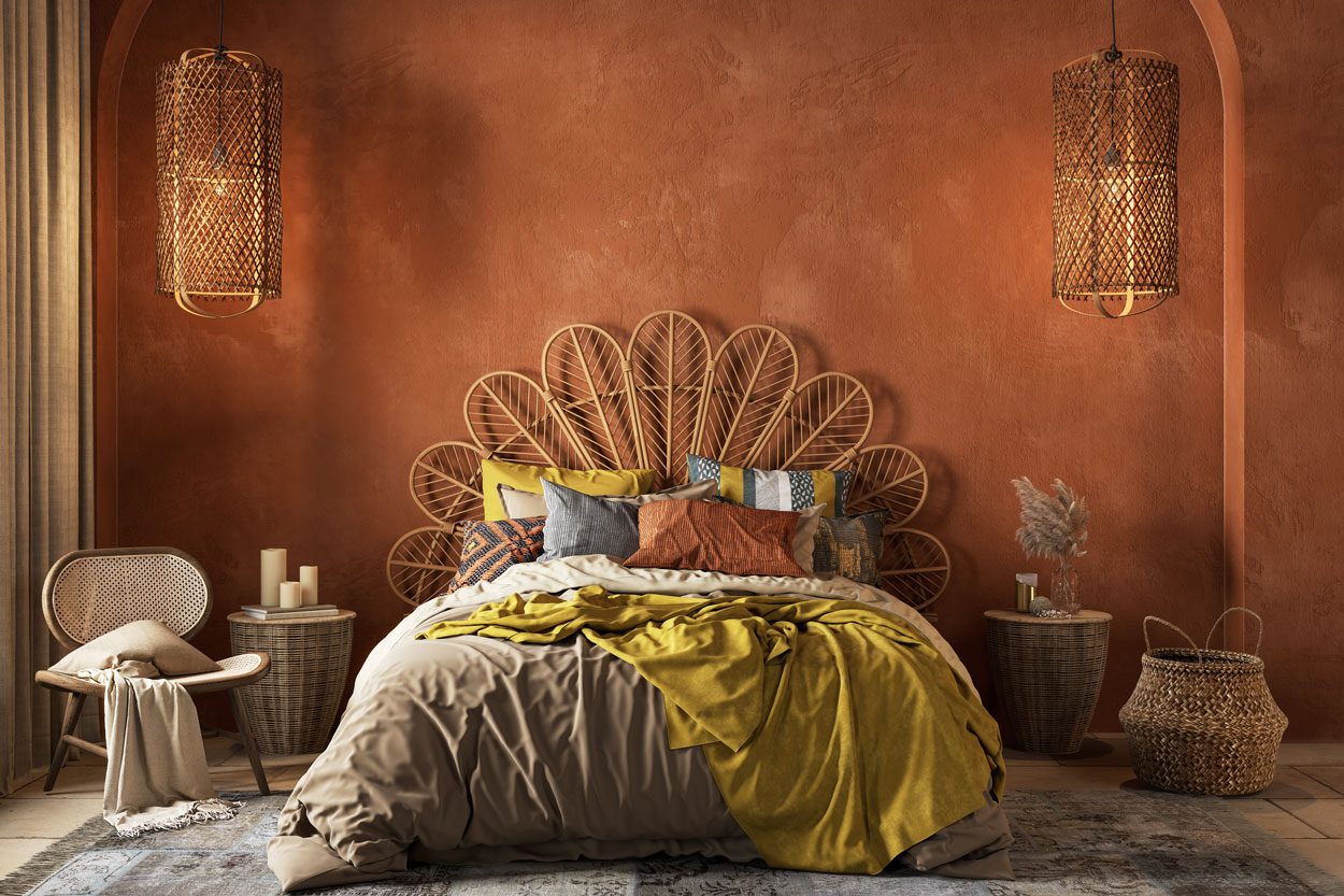 A cozy bed with colorful pillows and a blanket is situated in a warmly lit bedroom featuring an orange wall and woven decorations.