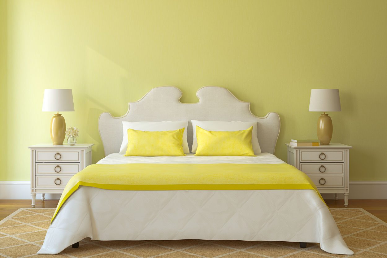 A bed with a curved headboard, yellow pillows, and a matching blanket is centered in a light yellow room with nightstands and lamps on either side.