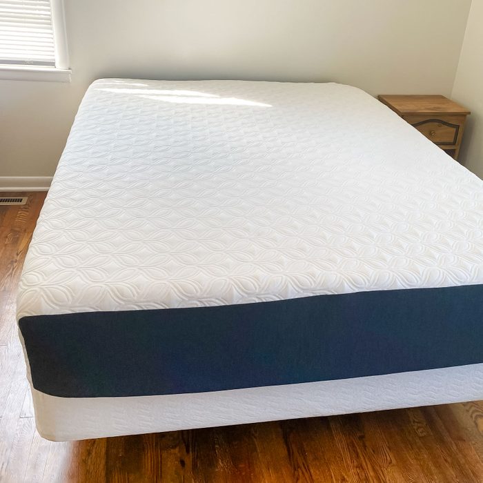 A neatly made bed with a patterned white mattress topper sits on polished wooden flooring, adjacent to a small wooden nightstand. Sunlight filters through a window.