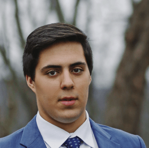 A man in a blue suit and tie stands outdoors, gazing directly at the camera, with blurred trees and a cloudy background behind him.