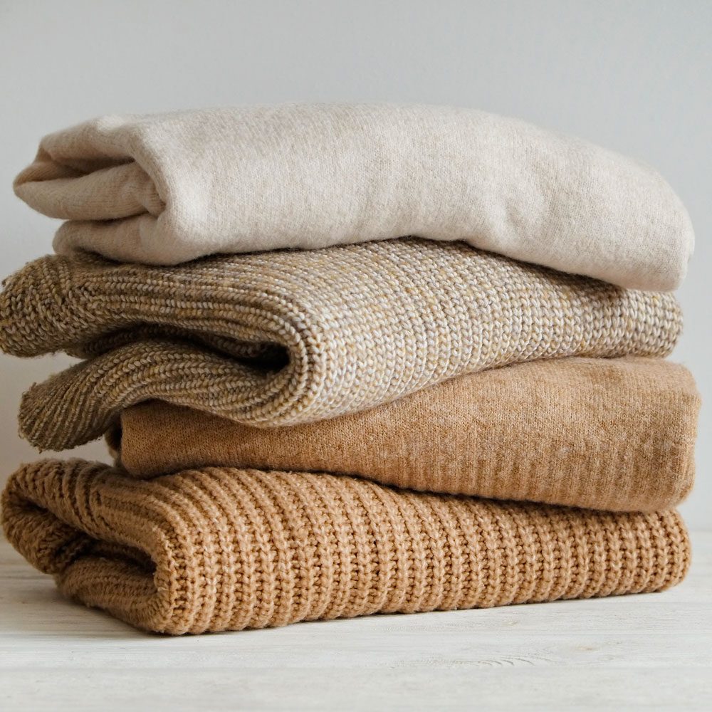 Stack Of Knitted Cashmere Sweaters stacked in a messy pile on a white wooden table and a light gray background