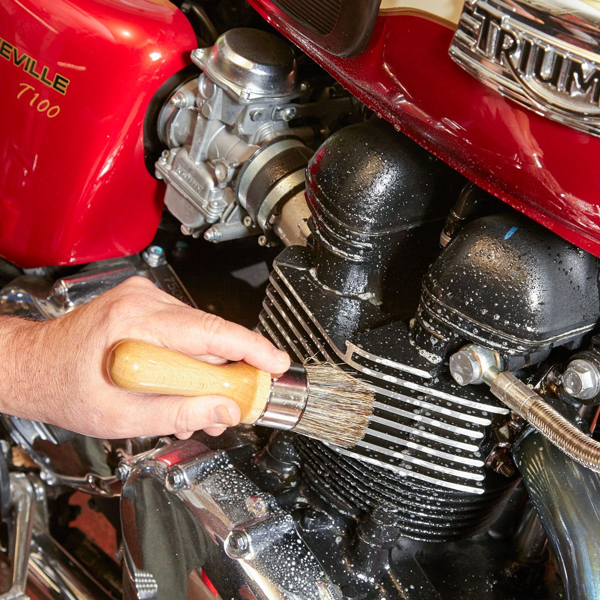A hand using a brush cleans the engine of a motorcycle, while soap bubbles and water are visible on the surface, revealing a polished chrome area.