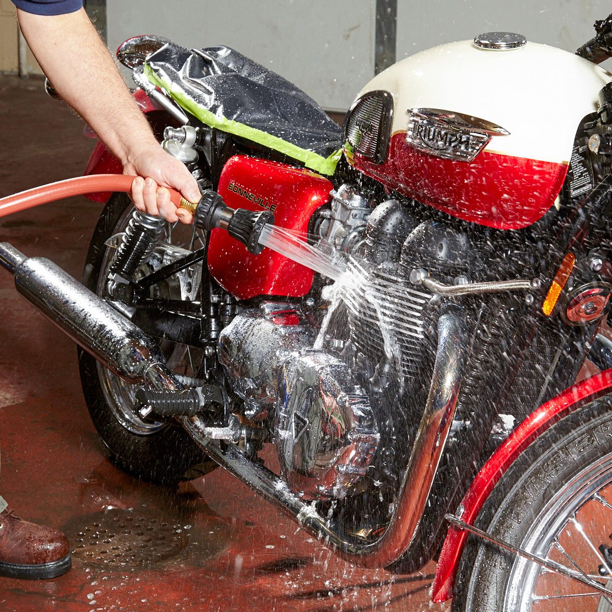 A person uses a hose to spray water on a motorcycle in a garage, cleaning its engine and body while water splashes around.