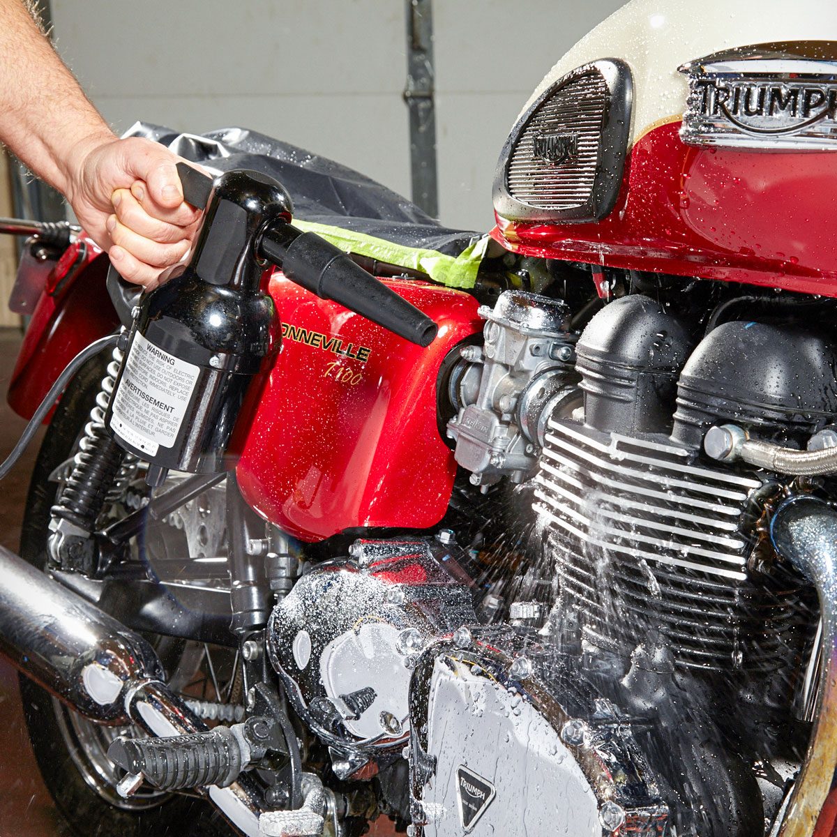 A person sprays water on a red motorcycle in a garage, cleaning it while droplets and foam fly off the surface.