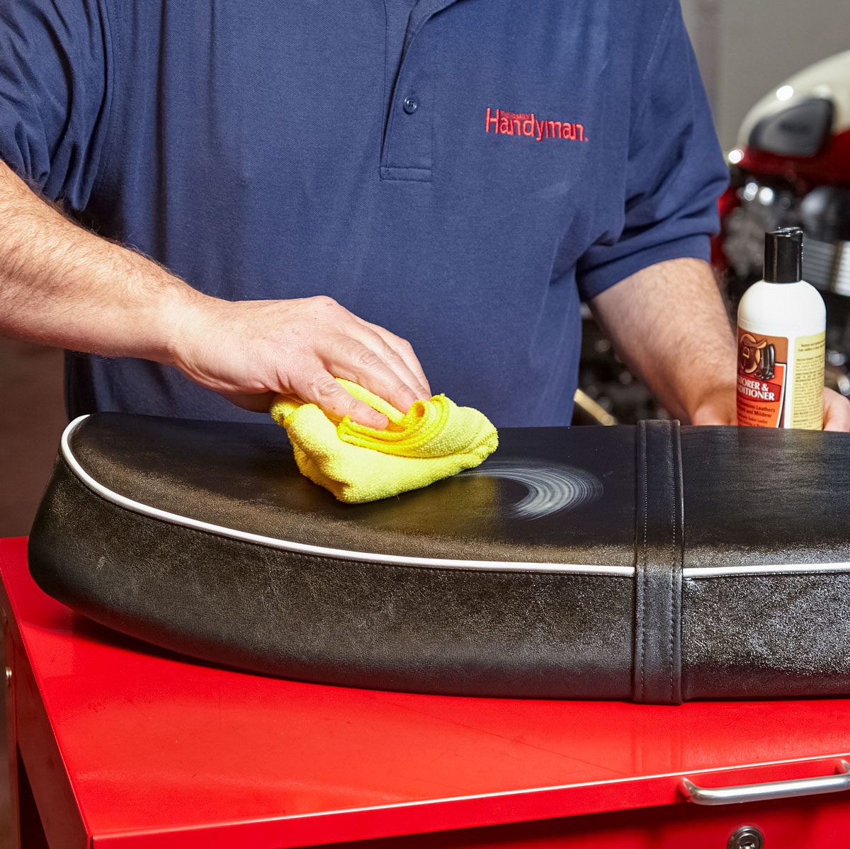 A person cleans a black leather motorcycle seat with a yellow cloth, using a bottle of conditioner on a red workbench in a garage setting.