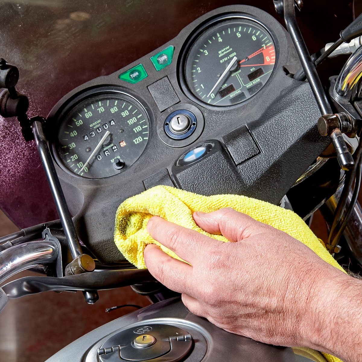 A hand wipes a motorcycle's dashboard with a yellow cloth, revealing gauges and controls, in a well-lit, indoor setting.
