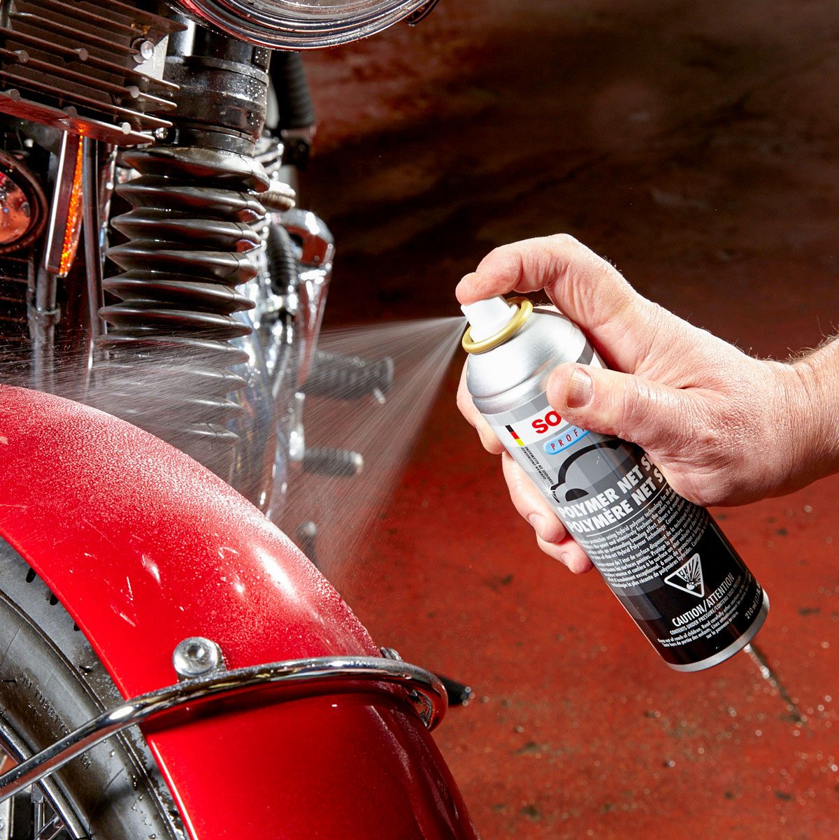 A hand sprays a liquid onto a motorcycle's red fender. The motorcycle is positioned on a textured surface with visible lighting, suggesting a maintenance setting.