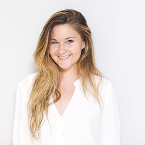 A woman with long, wavy hair smiles warmly while wearing a white shirt, standing against a light gray wall.
