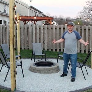 A man stands by a fire pit surrounded by chairs on a gravel circle, illuminated by string lights in a backyard setting.
