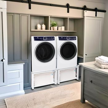 Two front-loading washing machines are placed side by side in a laundry room, surrounded by cabinetry, shelves, and a neatly folded stack of towels.