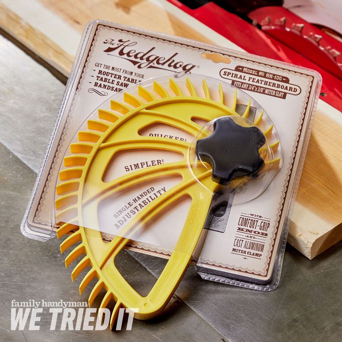 A yellow spiral featherboard rests on a wooden surface, packaged for use with power tools, labeled for features like adjustability and comfort grip.