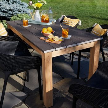 A wooden table showcases drinks and lemons, surrounded by black chairs, on a sunny outdoor patio with green grass and a blue tree backdrop.