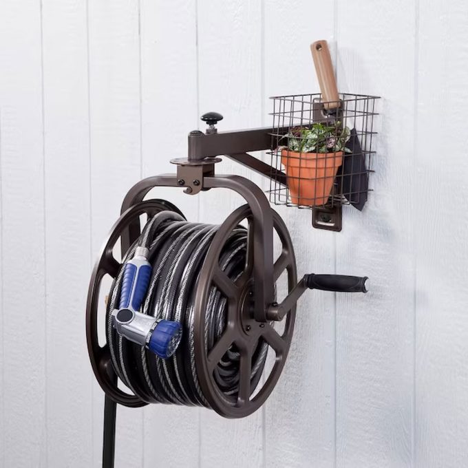 A wall-mounted hose reel holds a coiled garden hose with a spray nozzle, adjacent to a small planter in a wire basket against a wooden background.