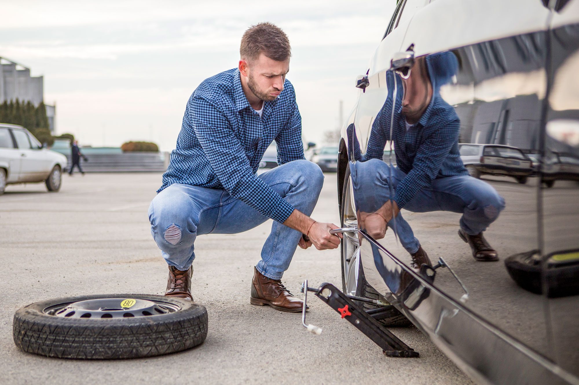 A man kneels by a car, using a wrench to change a tire, with a spare tire beside him in a parking lot.