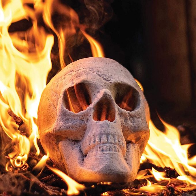 A white skull rests amidst flickering flames, with smoke swirling around in a dark, shadowy environment, creating an ominous and intense atmosphere.
