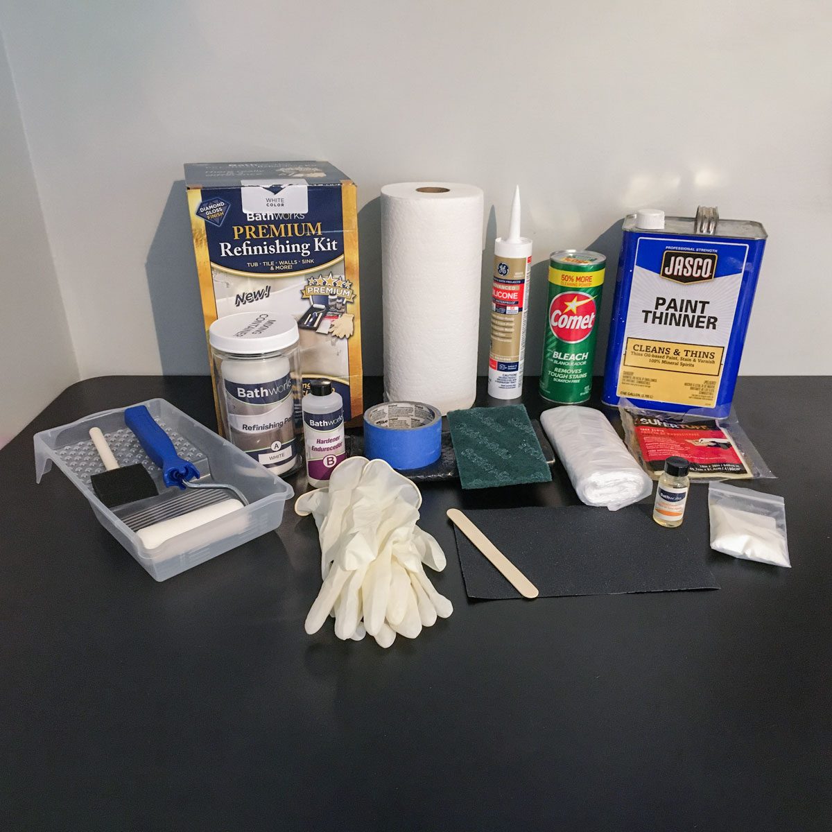 Various DIY supplies are arranged on a table, including a refinishing kit, cleaning agents, tools, and protective gear for a home improvement project.