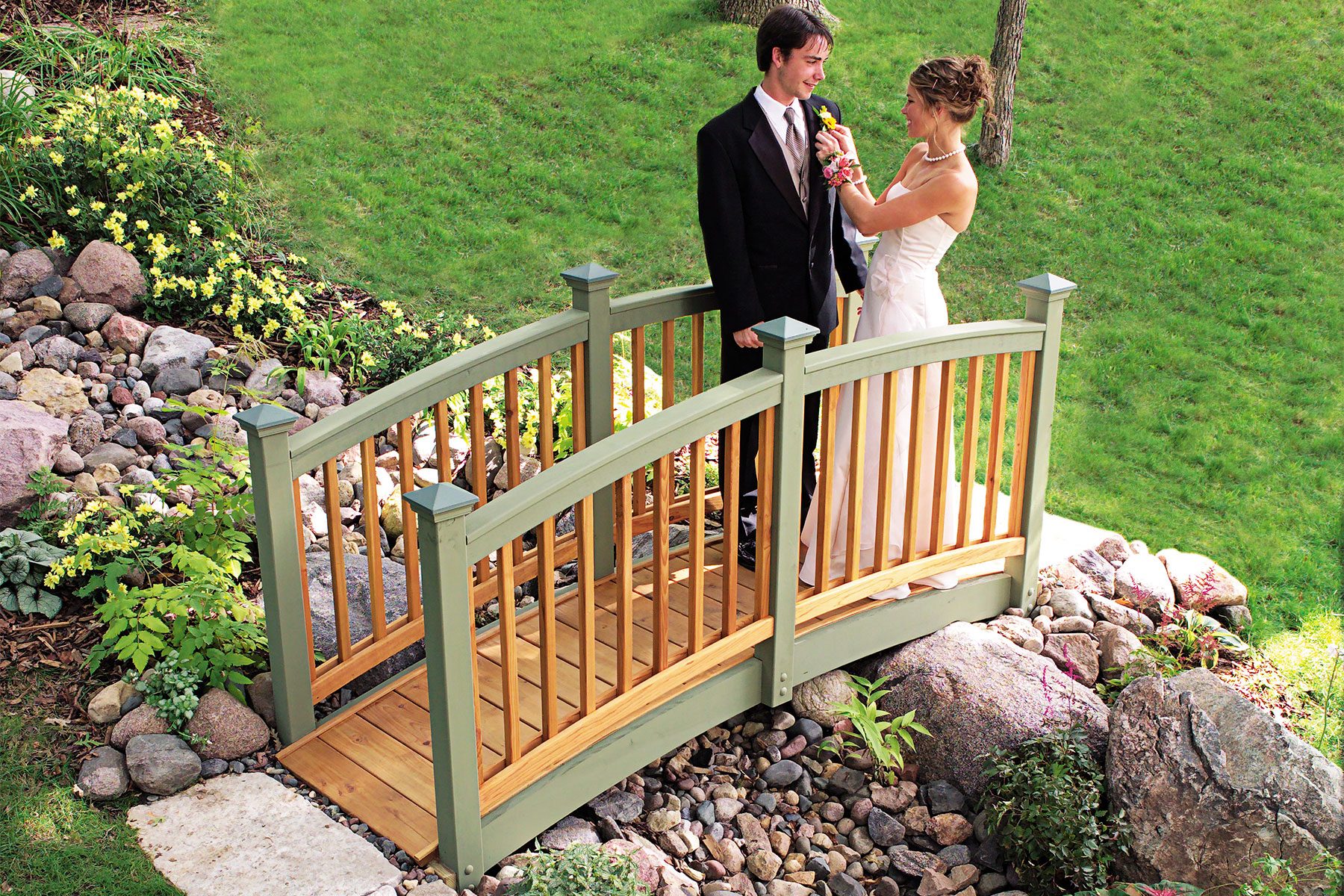 A bride and groom stand on a small wooden footbridge in a garden. The bride adjusts the groom's boutonniere while they smile at each other. The scene is surrounded by green grass, rocks, and various plants.