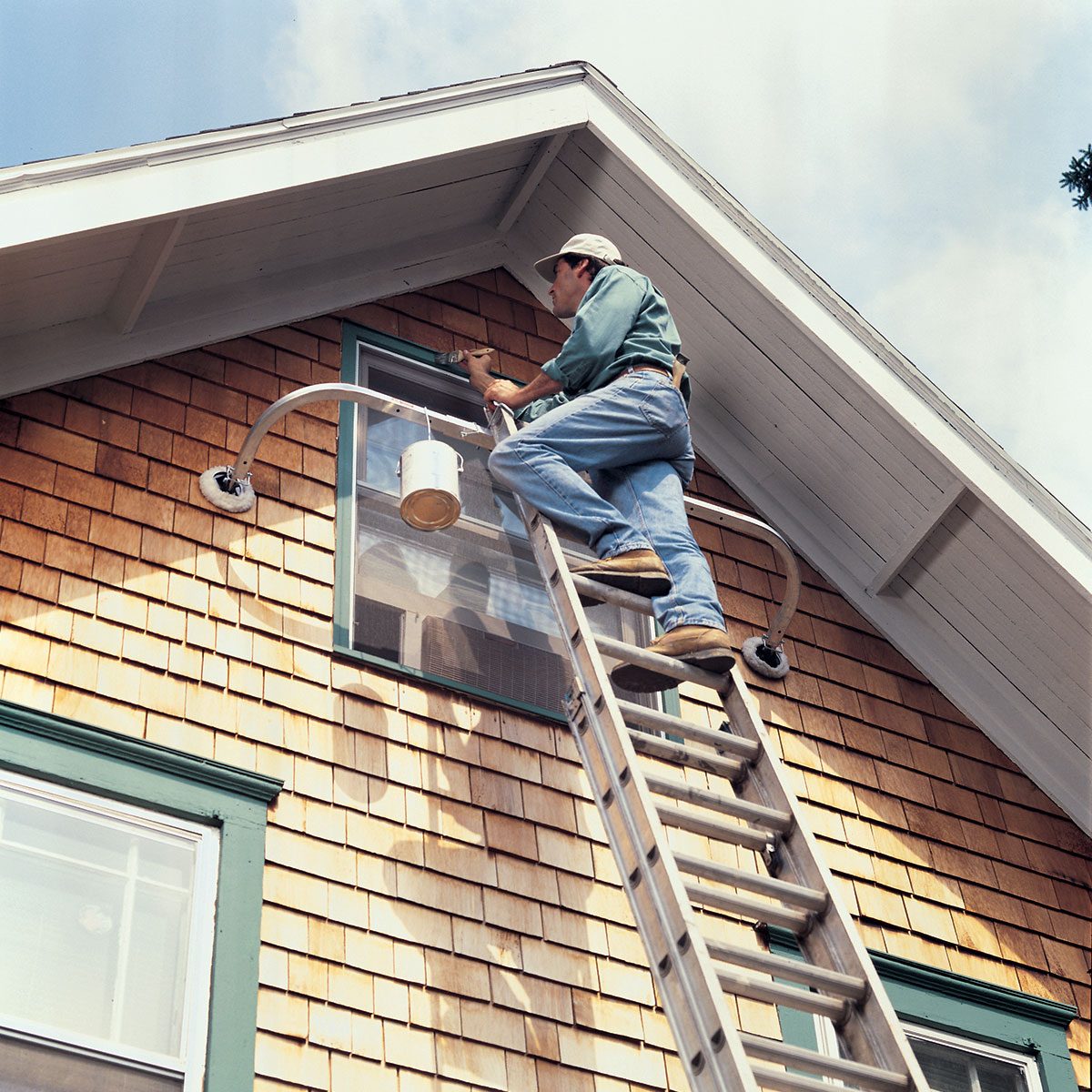 A person in a green shirt and jeans stands on a tall ladder, painting the upper exterior of a house with wood siding. They are using a paintbrush and a bucket of paint attached to the ladder. The house features a gable roof and green-trimmed windows.