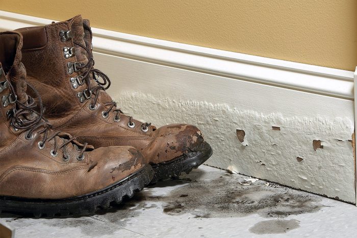 A pair of muddy brown work boots is positioned beside a baseboard with significant water damage. The baseboard paint is peeling and cracking, with visible signs of mold or dirt staining the floor below it. The wall has a tan color.