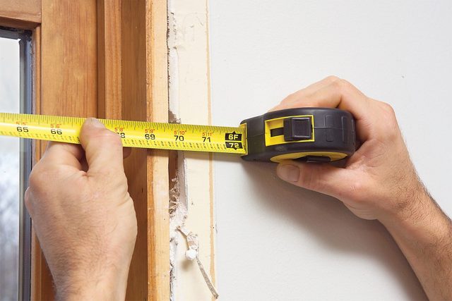 A person uses a yellow tape measure to measure the width of a window frame. One hand holds the tape dispenser, while the other extends the tape across the frame. The window has a wooden border and part of the bare wall is visible.