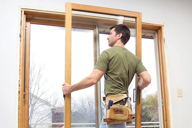 A man in a green shirt and tool belt is installing a sliding glass door. The door frame is wooden, and the man is positioned inside a house, working with his hands on the door. The background outside the door shows some trees and a cloudy sky.