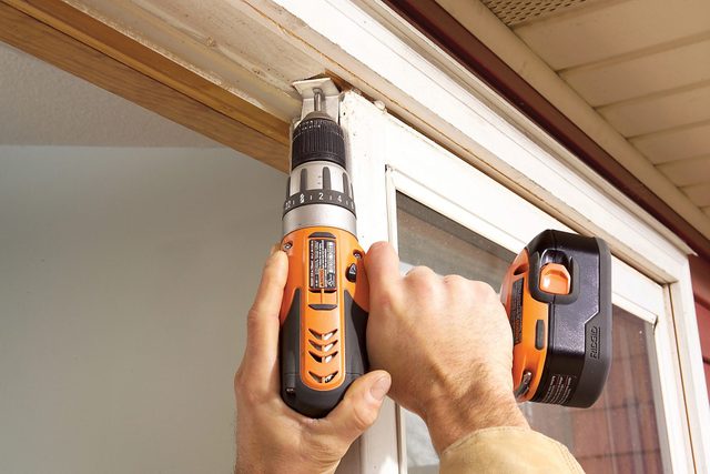 Close-up of hands using an orange and black power drill to fix a lock on a white door frame, with part of a house exterior visible in the background.