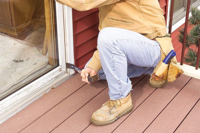 A person wearing light blue jeans, a brown jacket, and work boots is crouched near a sliding glass door, holding a tool and performing maintenance. The person has a tool belt with various tools attached, and the house has a red exterior with white trim.