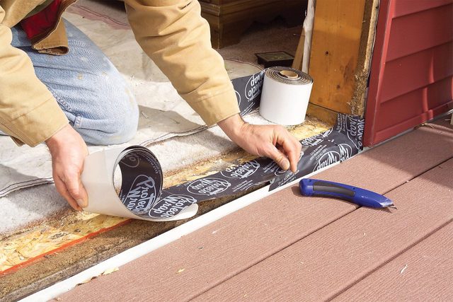 A person in a long-sleeve shirt is applying waterproof adhesive tape to a threshold area. There are two rolls of the tape nearby, and a blue-handled tool is on a wooden deck surface beside them. The scene appears to be part of a home improvement or construction project.