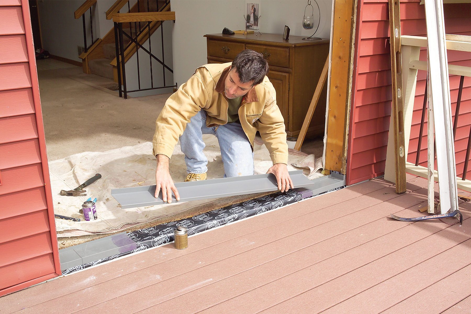 A person in a brown jacket installs a threshold on the floor where an interior room meets an exterior deck. Tools and materials are scattered around the work area, and the backside of wooden stairs is visible in the indoor space.