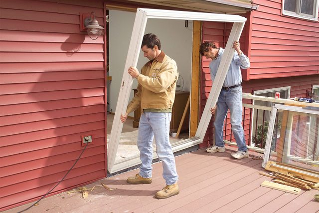 Two men are installing a white framed glass door in the doorway of a red house. They are working together to fit the doorframe into place. The house has red siding and a wooden deck. Various tools and materials are scattered around them.