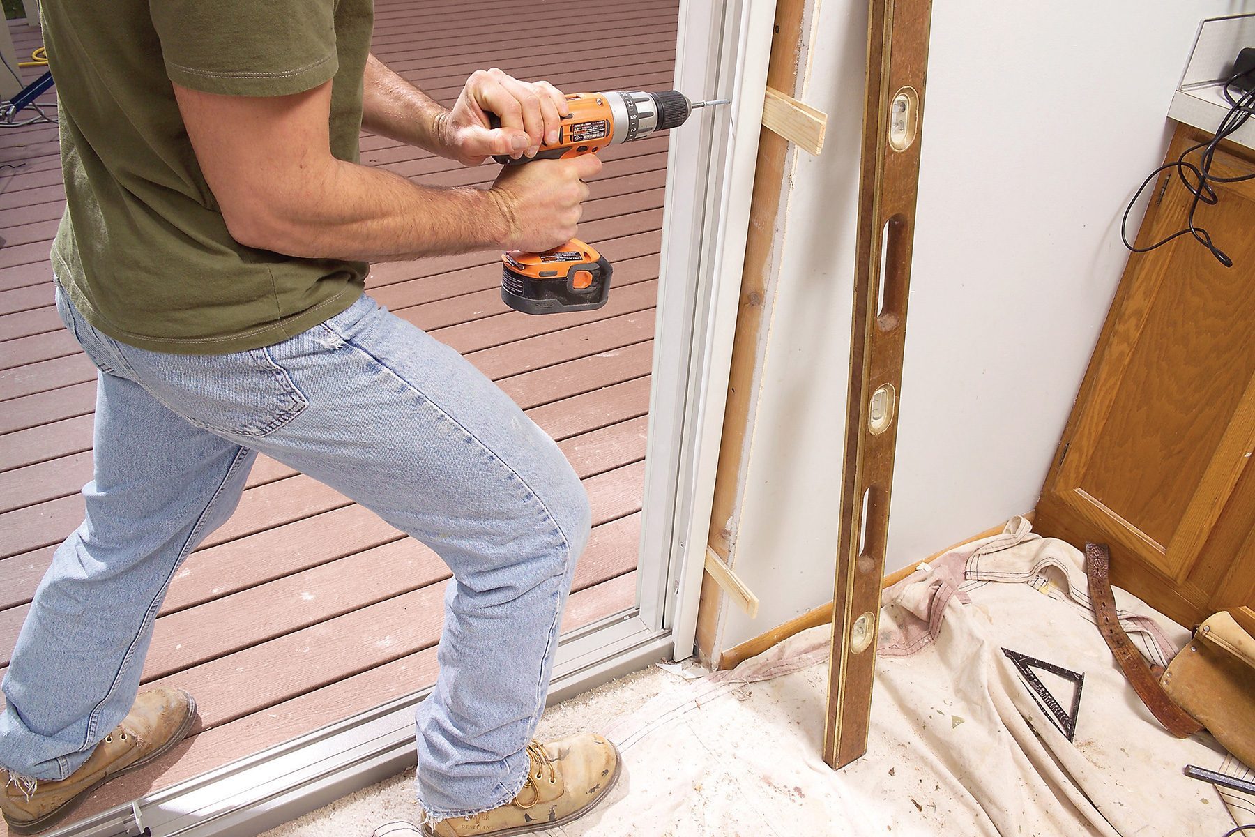 A person wearing a green shirt, blue jeans, and tan work boots is using an orange cordless drill to install a door frame. A wooden level is leaning against the wall beside the person. The floor is covered with a dust sheet.