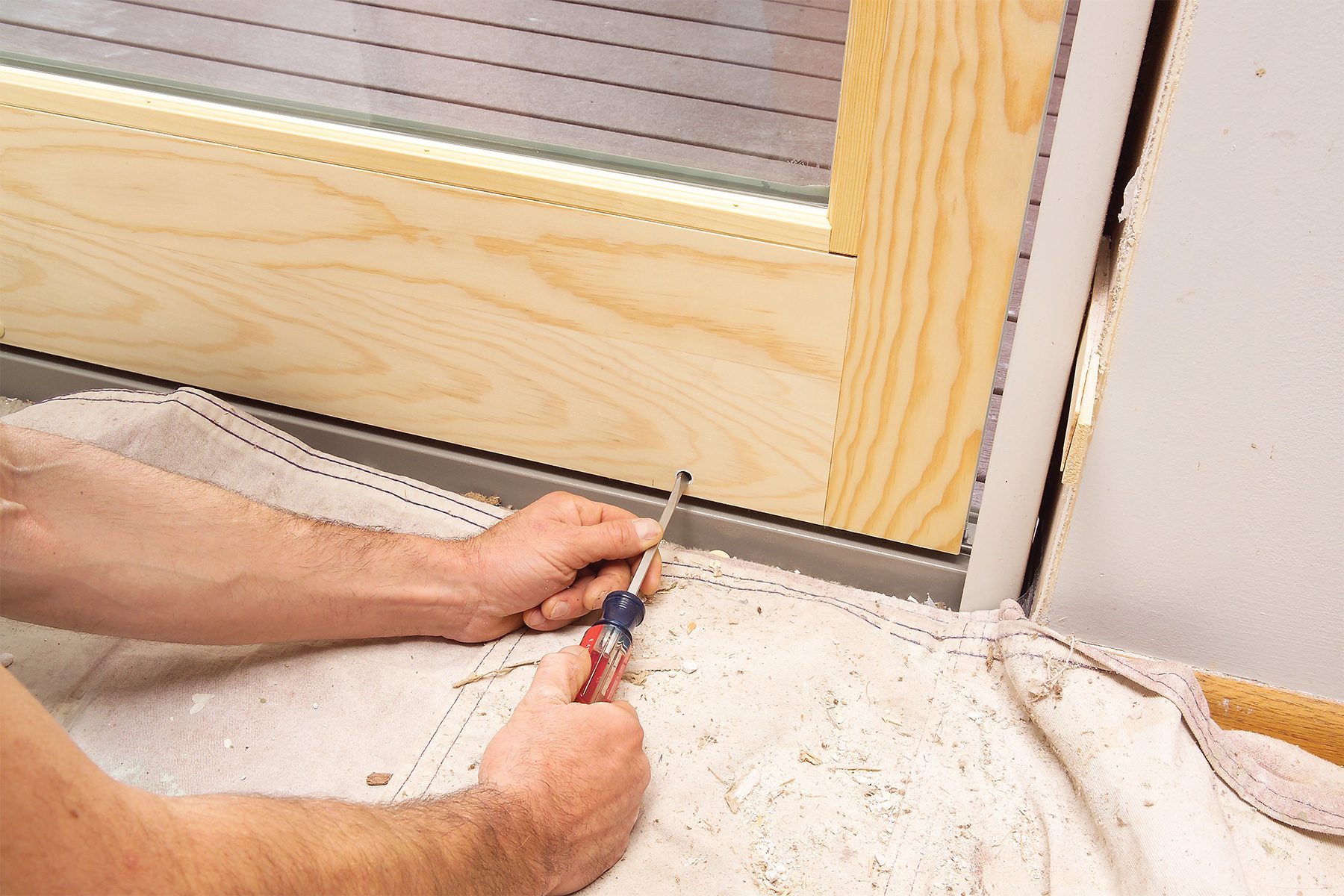 A person is using a flathead screwdriver to install or adjust a wooden door frame. The bottom part of the door and frame are visible, along with the person’s hands holding the tool. There is a cloth covering the floor to protect from dust and debris.