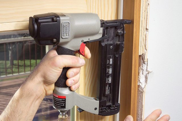 A person using a pneumatic nail gun to secure wooden trim on a window frame.