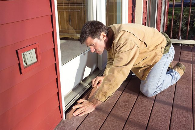 A man in a tan jacket and jeans installs weather stripping at the bottom of a red exterior door, kneeling on a wooden deck. The door is open, and the man is focused on placing the stripping correctly.