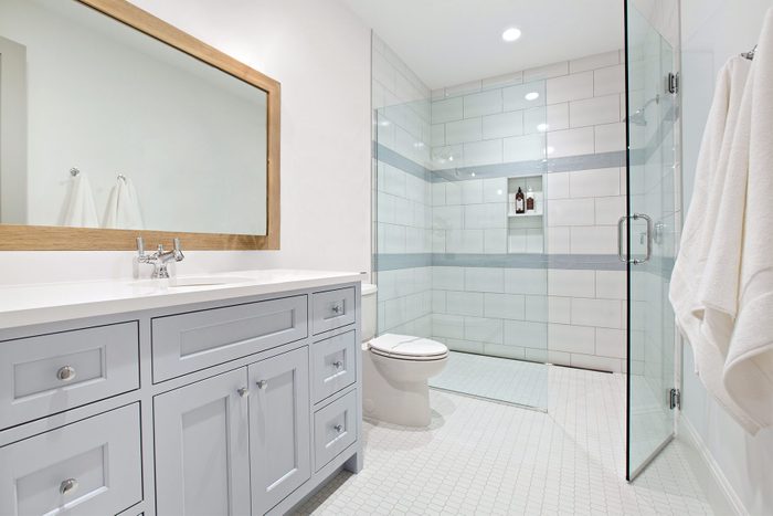 A modern bathroom featuring a white vanity with a wooden-framed mirror above it. There is a glass-enclosed walk-in shower with white and light gray tile. A white toilet is placed next to the shower. The floor is covered with small white tiles.