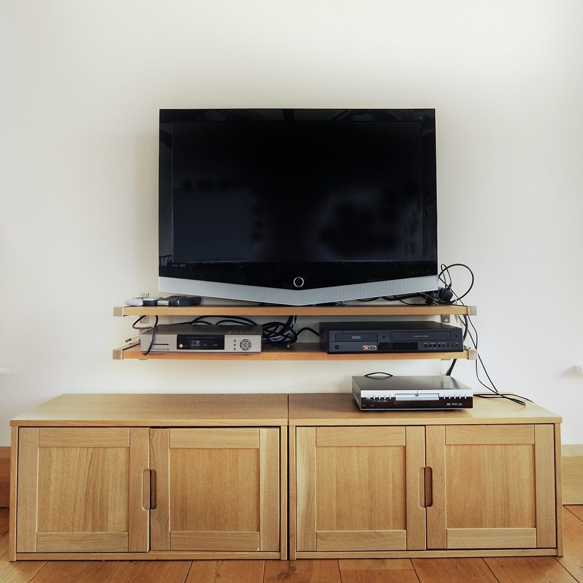A flat-screen TV is mounted on a wall above a wooden media console with two shelves. The shelves hold various electronic devices, including a satellite receiver, a router, and a gaming console. The console below has four closed cabinet doors. Cords are visible and untangled.