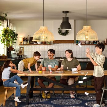 A family of five is gathered around a wooden dining table in a cozy kitchen. The parents are seated, smiling, while their three children are playfully interacting. Paper planes are flying, and the room is warmly lit with hanging wicker lamps.