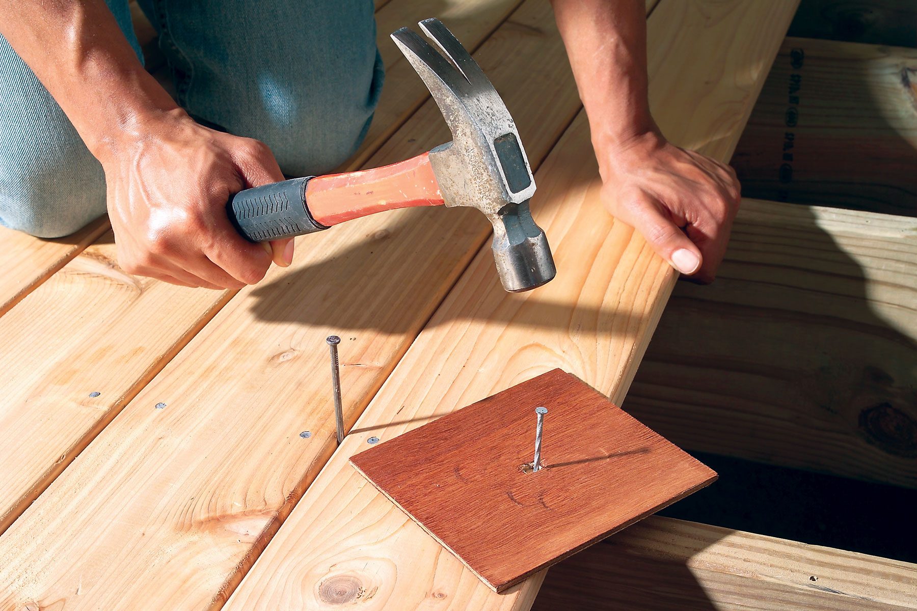 A person hammers a large nail into a wooden plank on a deck. They use a small square piece of wood to protect the plank