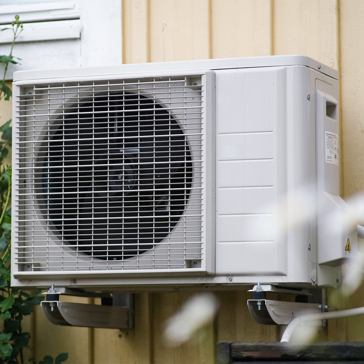 Outdoor HVAC unit mounted on the side of a yellow building. The unit has a large, circular fan with a protective grille on the front. Green foliage partially surrounds the bottom left corner of the unit.