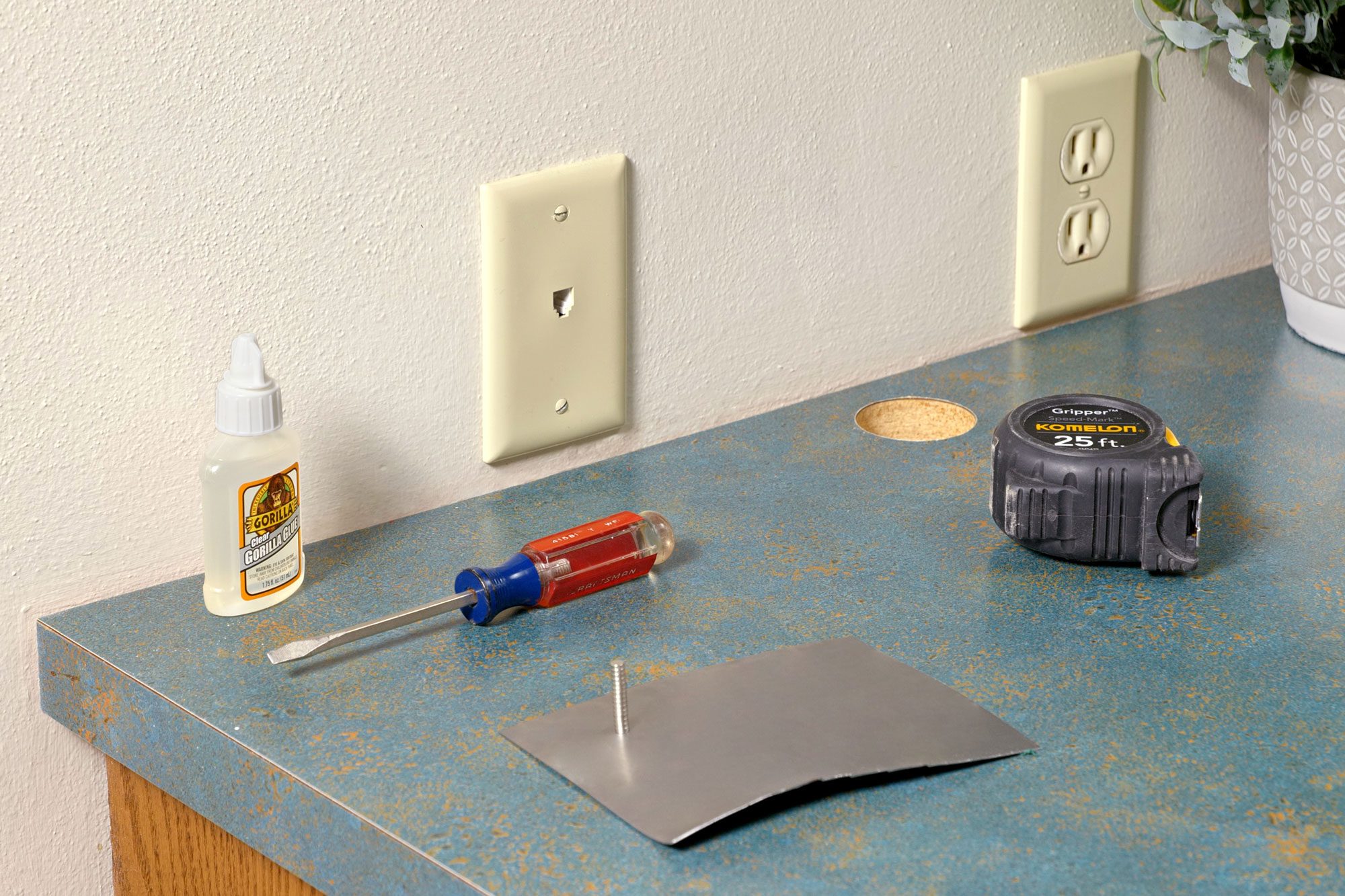A metal plate is placed on the surface, ready to be mounted or attached; Nearby, essential tools like a screwdriver and a measuring tape are laid out; A bottle of glue and a nail are also available for securing the plate; The countertop features two electrical outlets and a small potted plant;