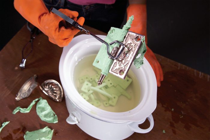 A person wearing orange gloves uses a pair of tongs to hold a vintage beater coated in green residue above a white container filled with liquid. Other beaters are on the table, showing similar green residue.