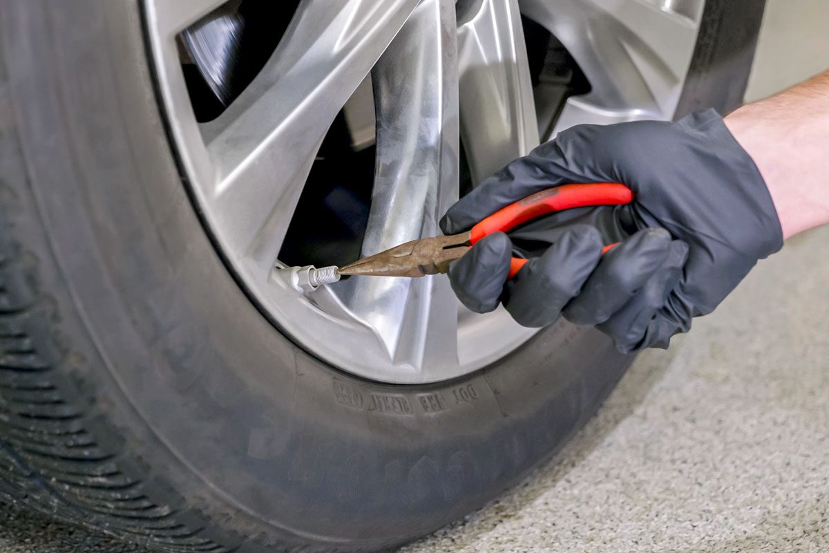 A person wearing black gloves uses pliers to remove a tire valve stem from car's tyre
