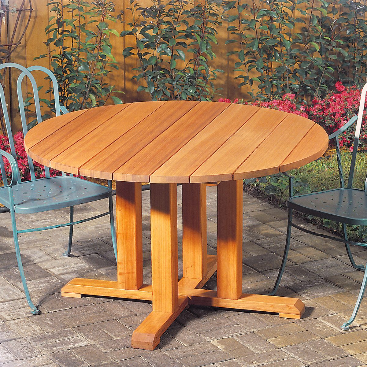 A round wooden table with a slatted top sits on a paved patio. Two metal chairs with curved backs are placed around it. In the background, there are green plants and red flowers along a wooden fence.