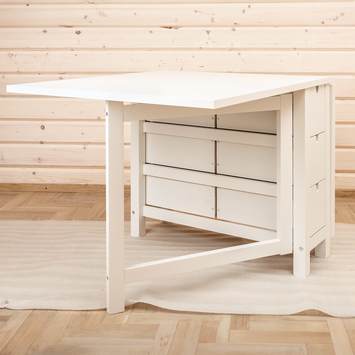 White drop-leaf table with drawers in front of a light wooden paneled wall. The table is placed on a light-colored checkered rug over a parquet floor.