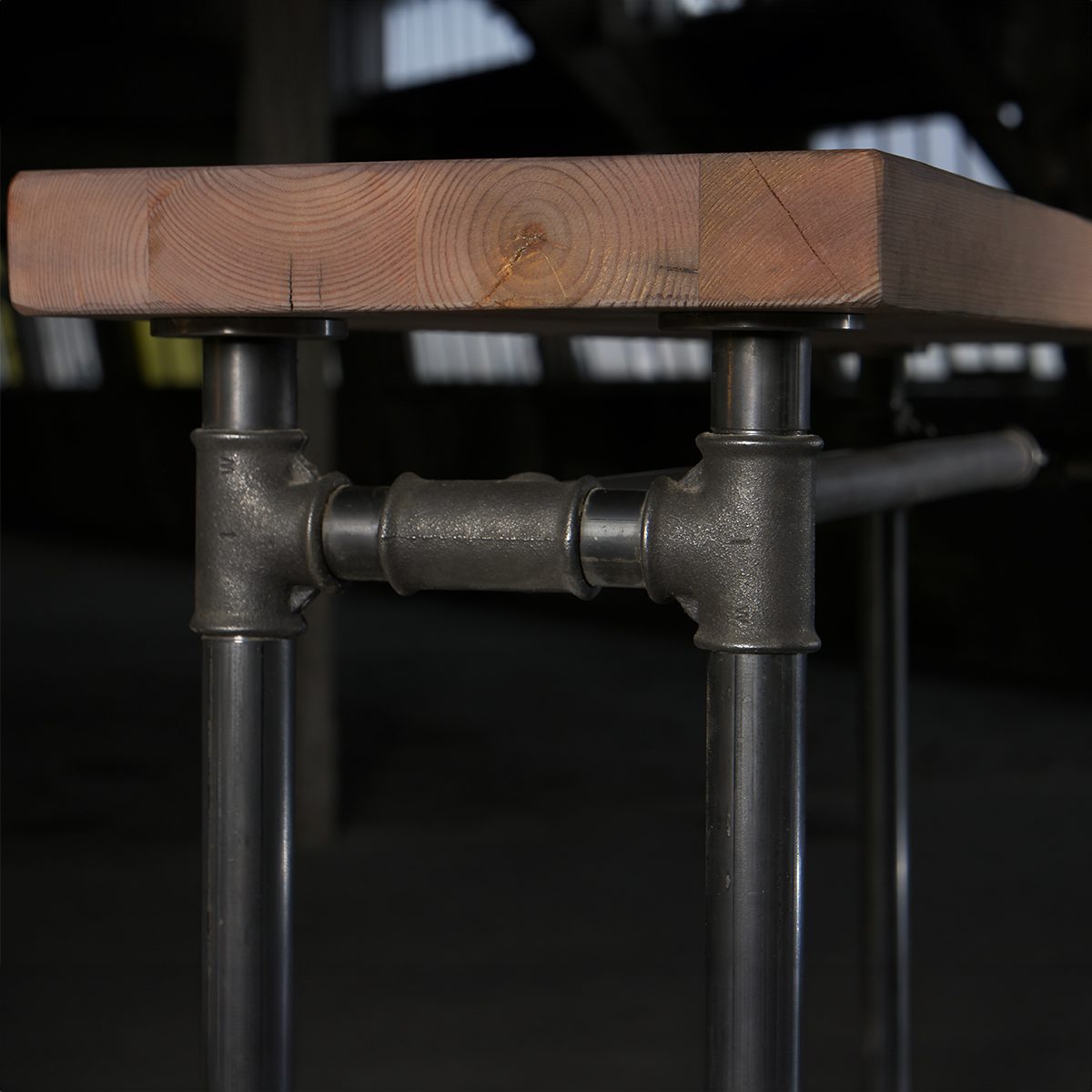 A wooden tabletop with visible rings is supported by industrial-style metal pipe legs and connectors in a dimly lit room.