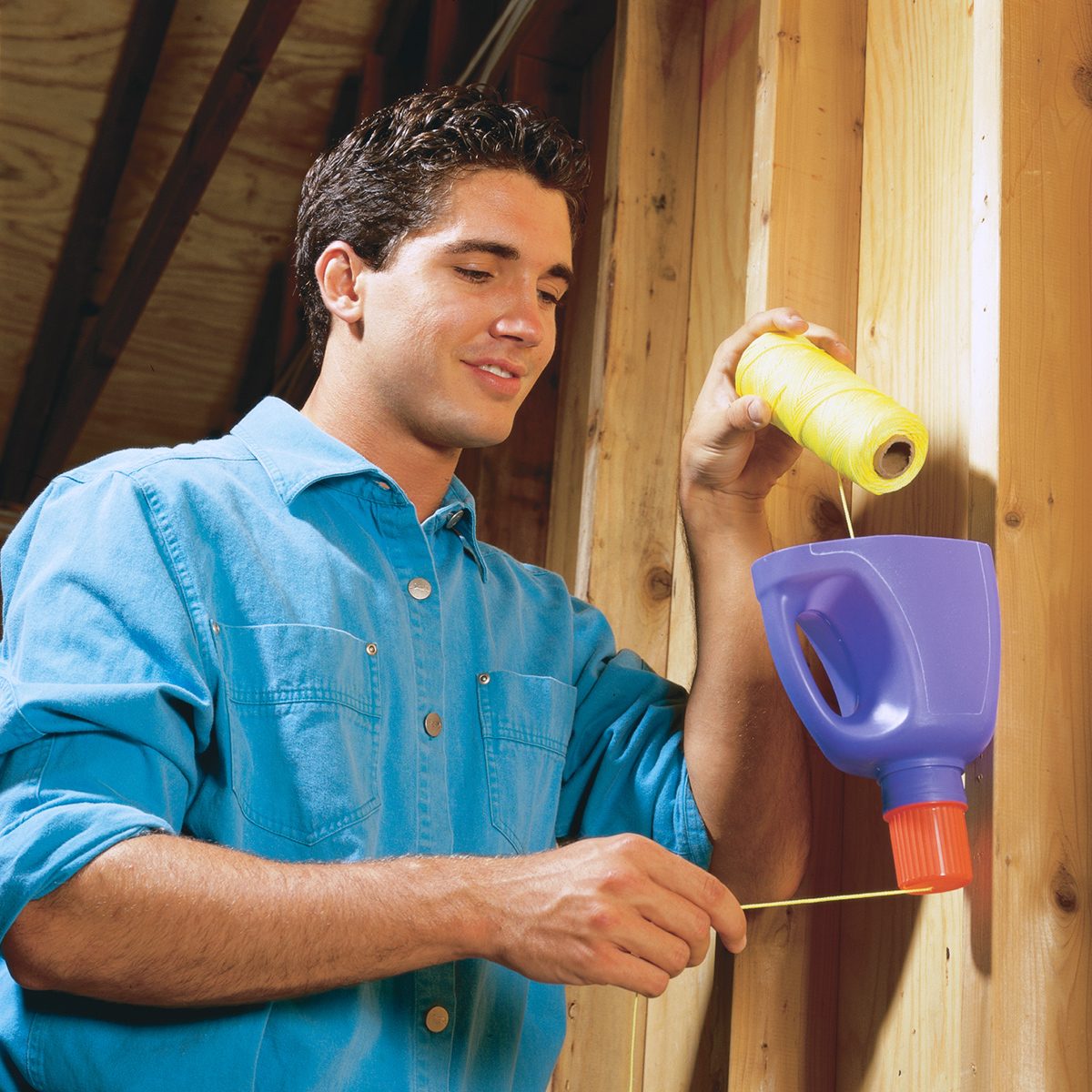 A person in a blue shirt is using a chalk line tool, pouring chalk powder into a blue and purple container. The tool is pressed against a wooden wall, and the person is pulling the string out for marking.