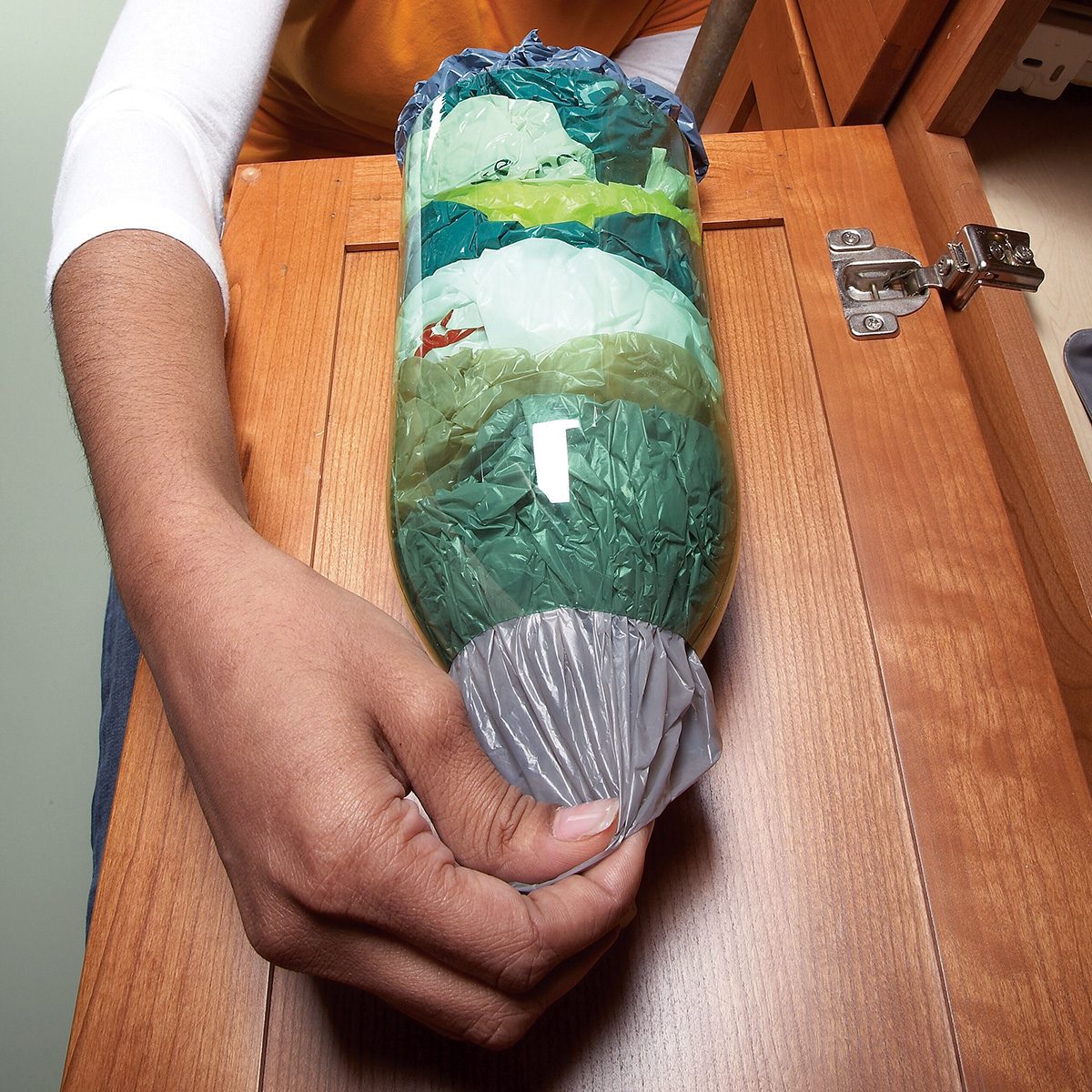A person holding a repurposed plastic bottle filled with colorful plastic bags, likely used as a bag storage dispenser. The bottle is cut and stuffed with bags in shades of green, blue, and red. Cabinet hinges are visible in the background.