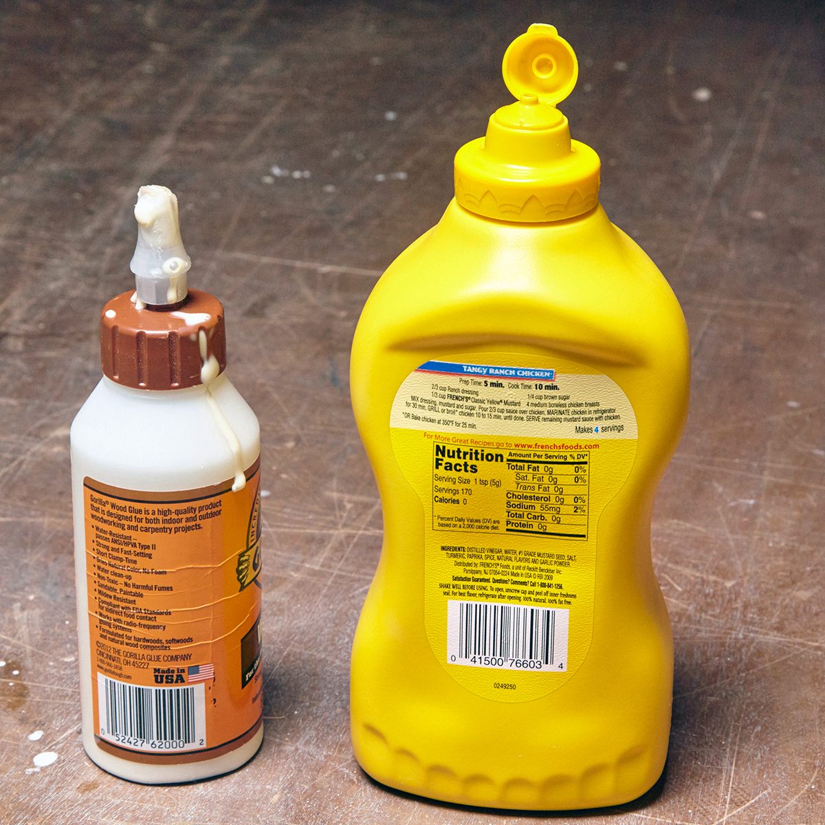Two condiment bottles on a tabletop: one small with a brown cap, and a larger yellow mustard bottle with an open flip-top lid. Both have labels with text and barcodes on them.