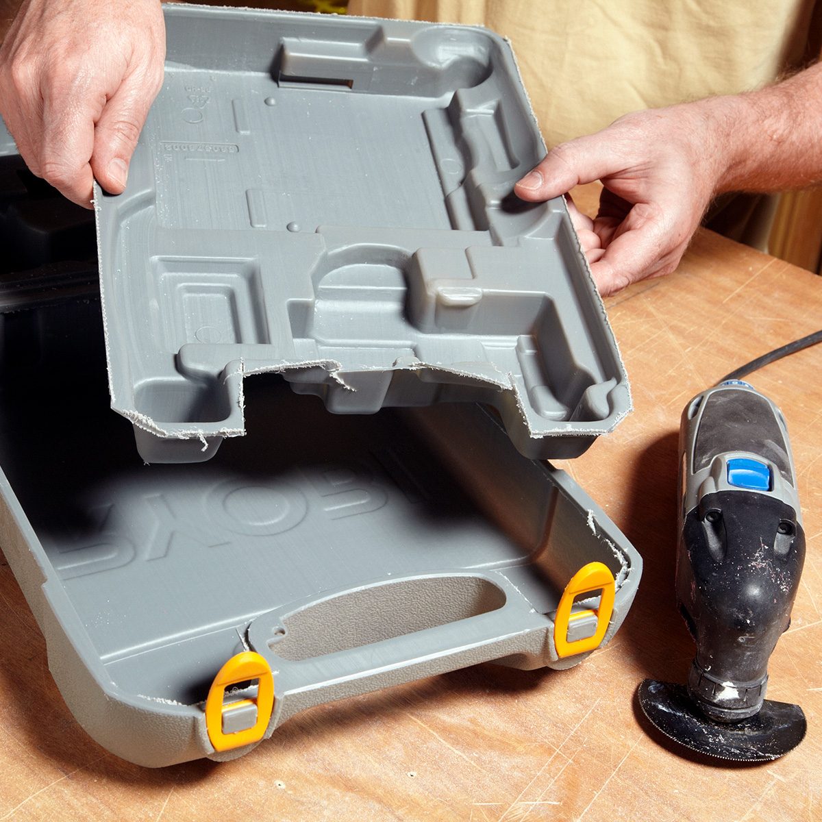 A person holds the open lid of a gray plastic toolbox with yellow latches on a wooden surface. An oscillating multi-tool is placed beside the toolbox.